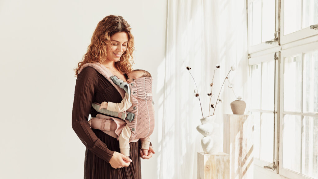 אמא נושאת תינוק במנשא מדגם בייבי ביורן הרמוני בצבע ורוד, בד אייר מש.
