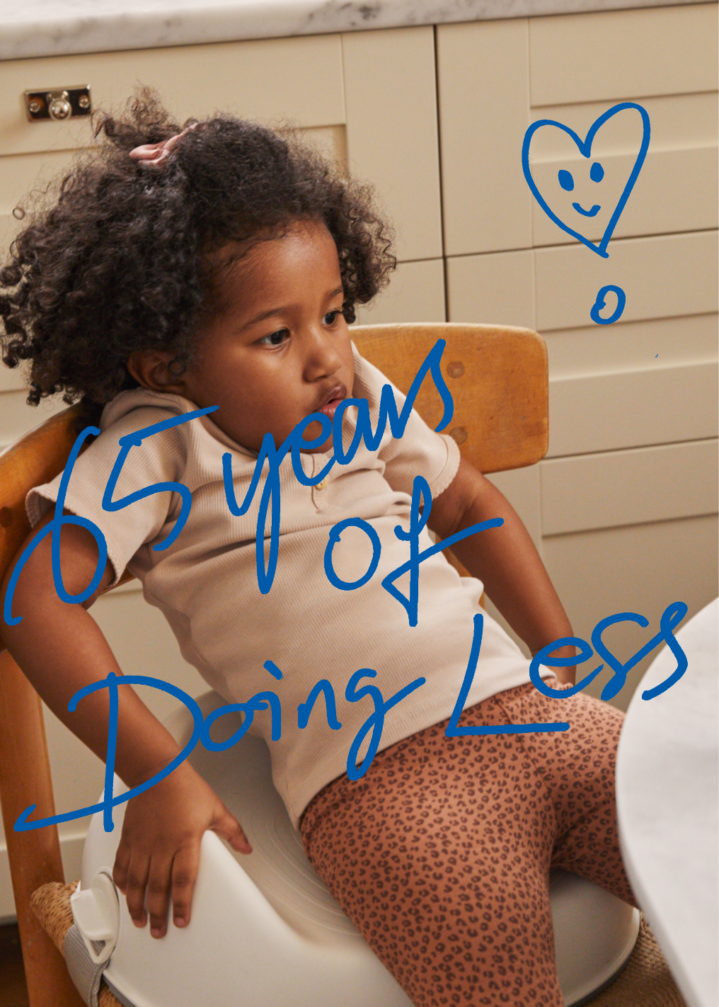 ילדה יושבת על בוסטר האכלה מול שולחן. כיתוב בכחול על גבי התמונה:
65 שנים של "לעשות פחות"